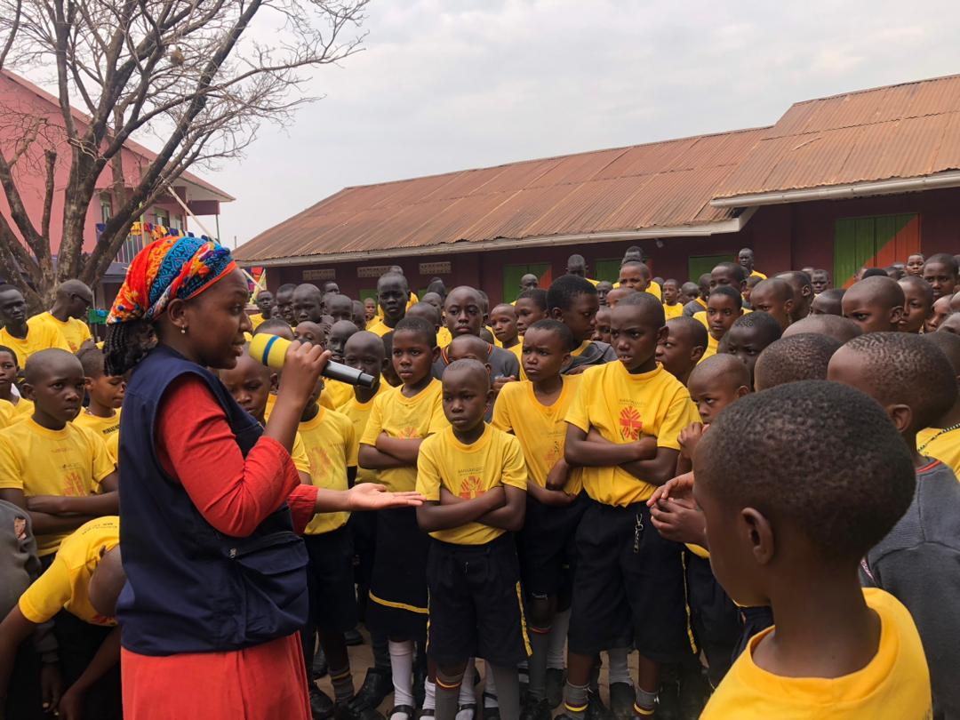 A facilitator addresses learners in yellow uniforms at an outdoor school gathering.